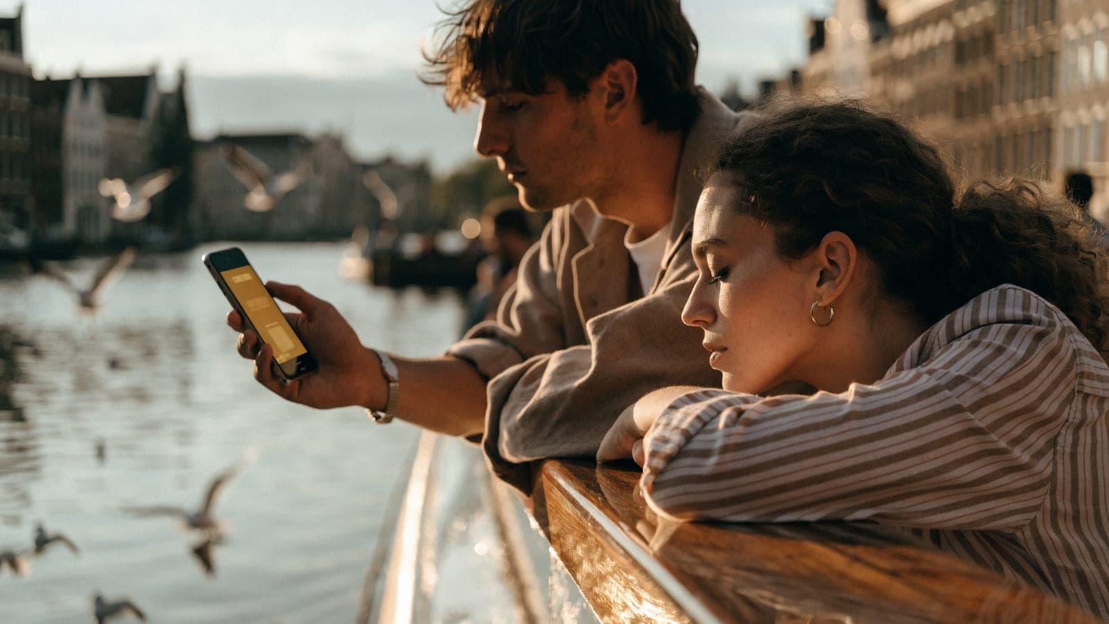 A couple at the railing of a river cruise ship at golden hour as a canal city comes into view, a phone showing a golden ticket